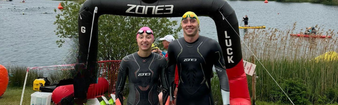 a man and a woman are facing the camera, both stood next to the start line the swim start of a triathlon wearing their wetsuits and brigh colpured swim caps