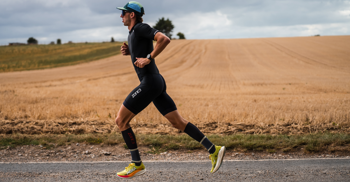 Man running on a road wearing zone3 trisuit with a field and trees in the background