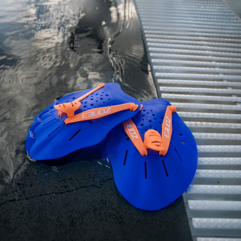 Pair of blue Ergo Swim Training Hand Paddles with orange straps resting on the pool edge beside a metal grate.