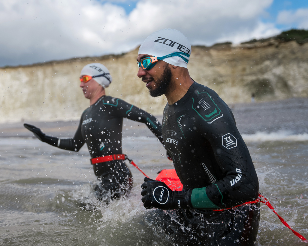 Two athletes in vision wetsuits and swim caps emerging from water with cliffs in the background