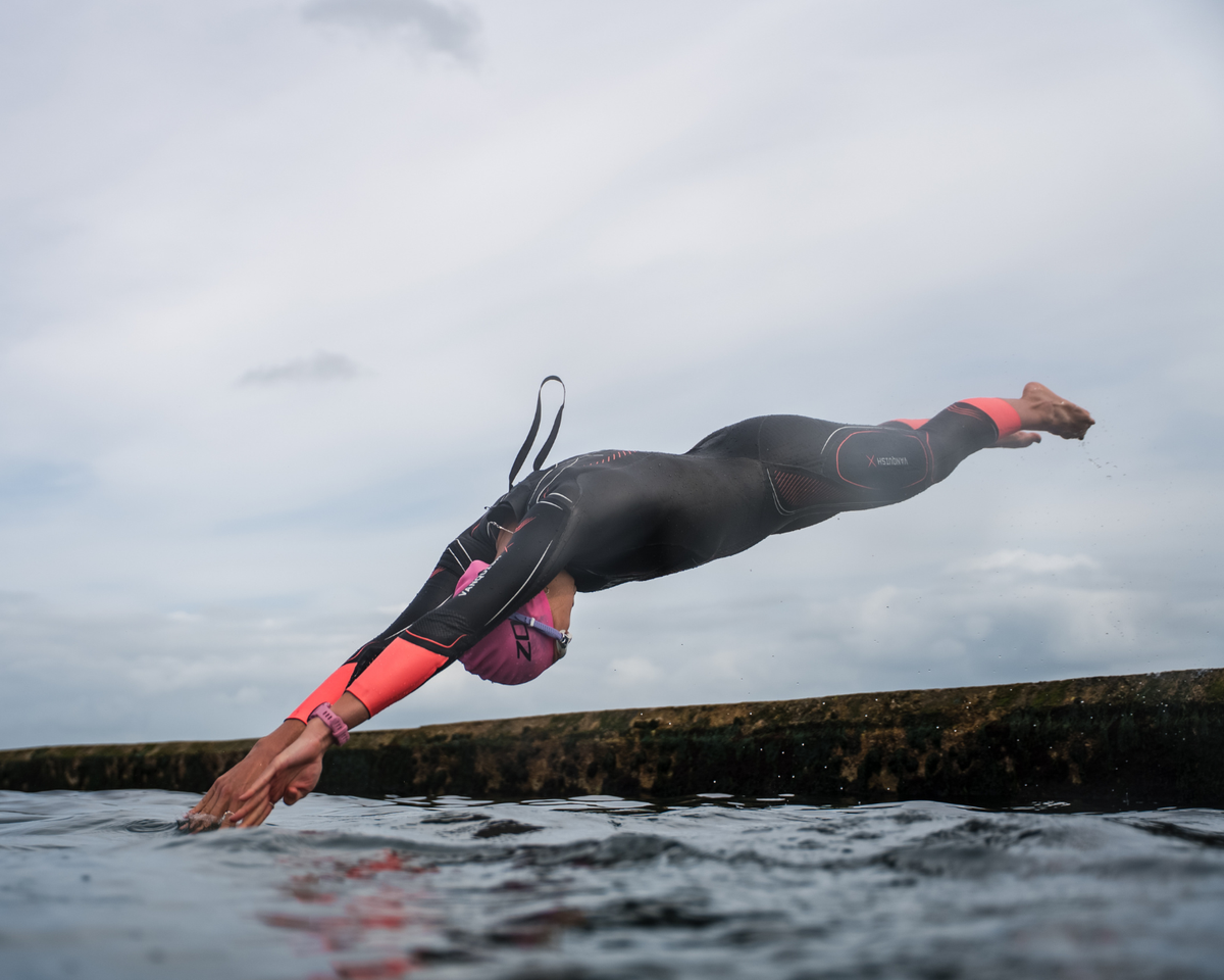 Person in Zone3 Vanquish-x wetsuit diving into water with a cloudy sky in the background