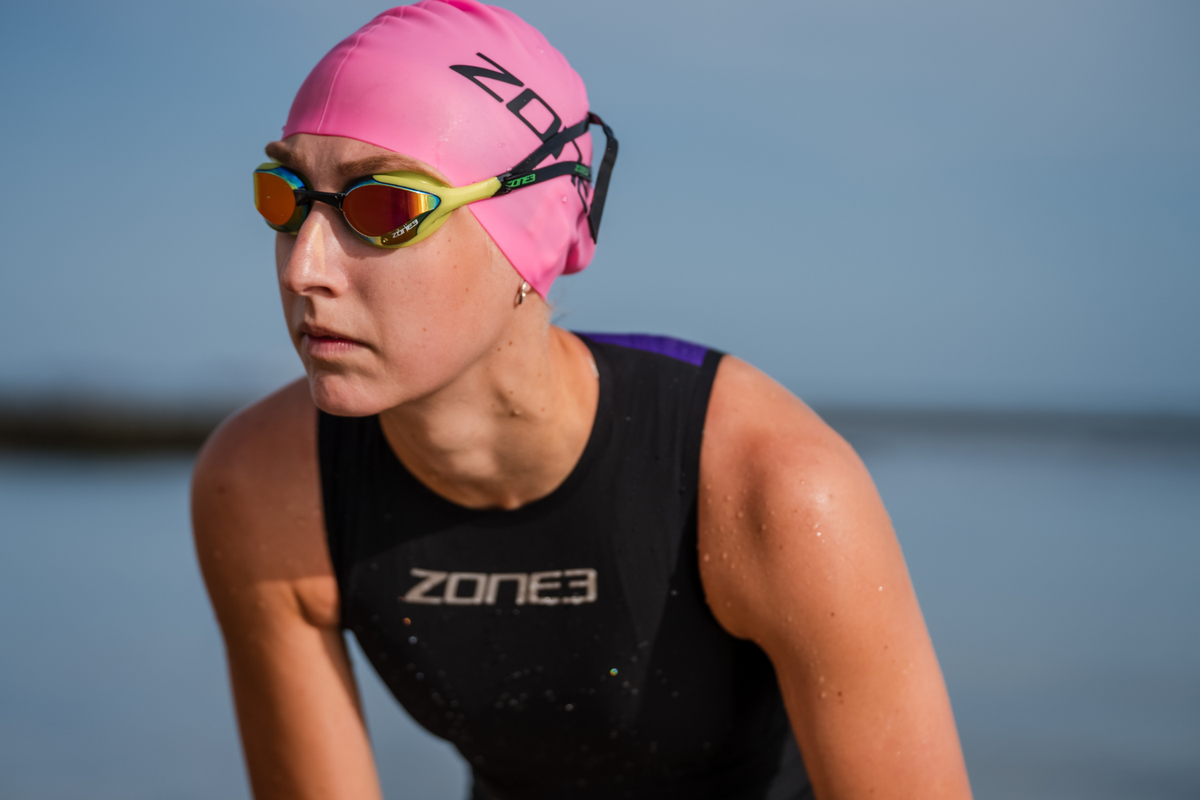 Person wearing a pink swim cap, racing swim goggles and a black sleeveless swimsuit. They are standing by a body of water.