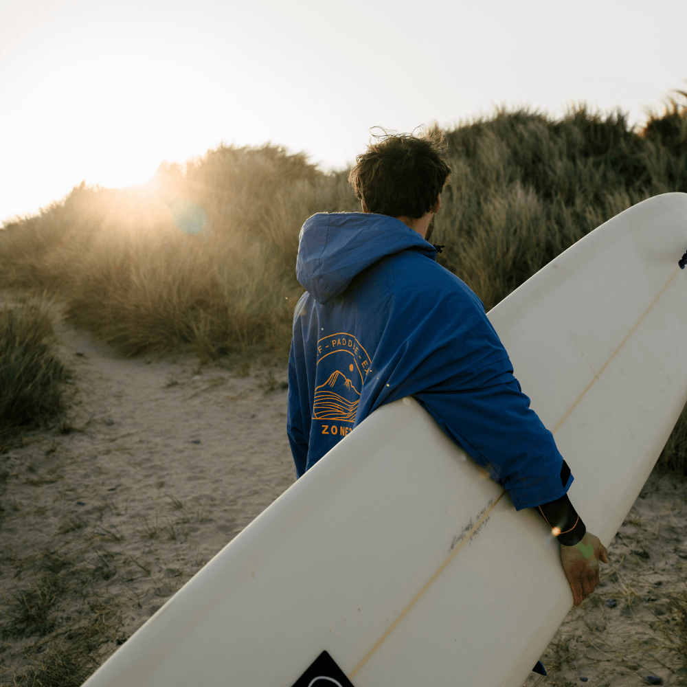 Rear shot of a person walking up a beach path at sunset, carrying a surfboard while wearing the ZONE3 robe.