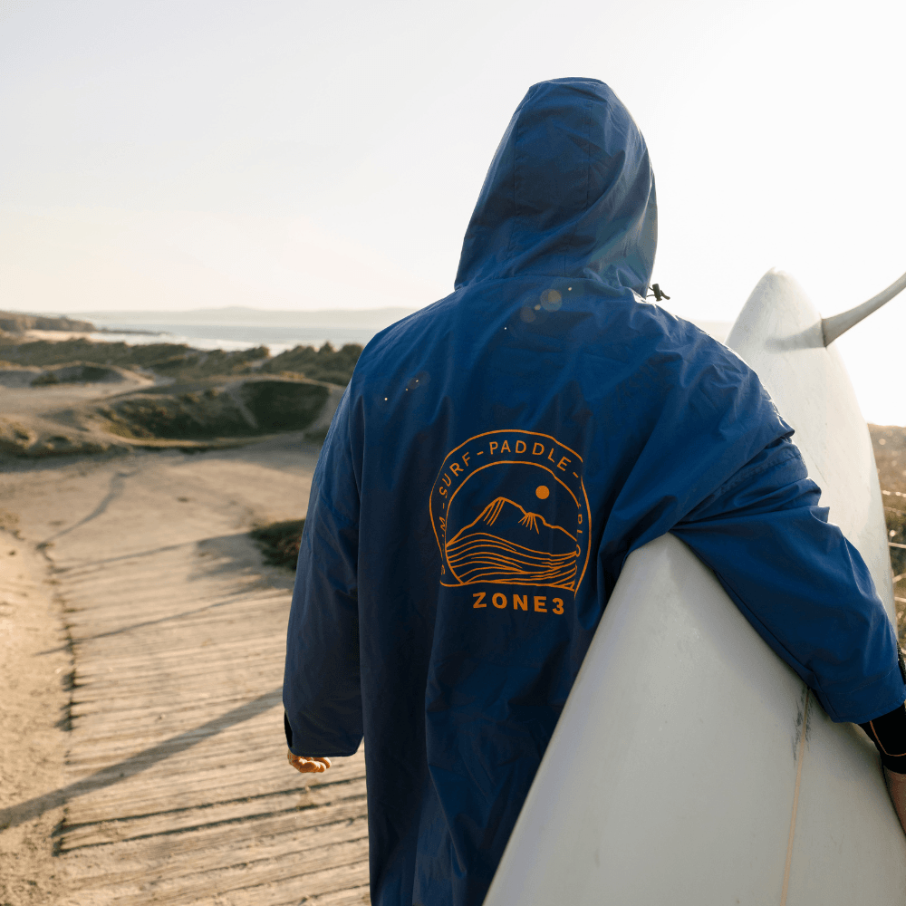 Person in a blue ZONE3 robe with hood up, walking with a surfboard under arm, large logo on back visible.